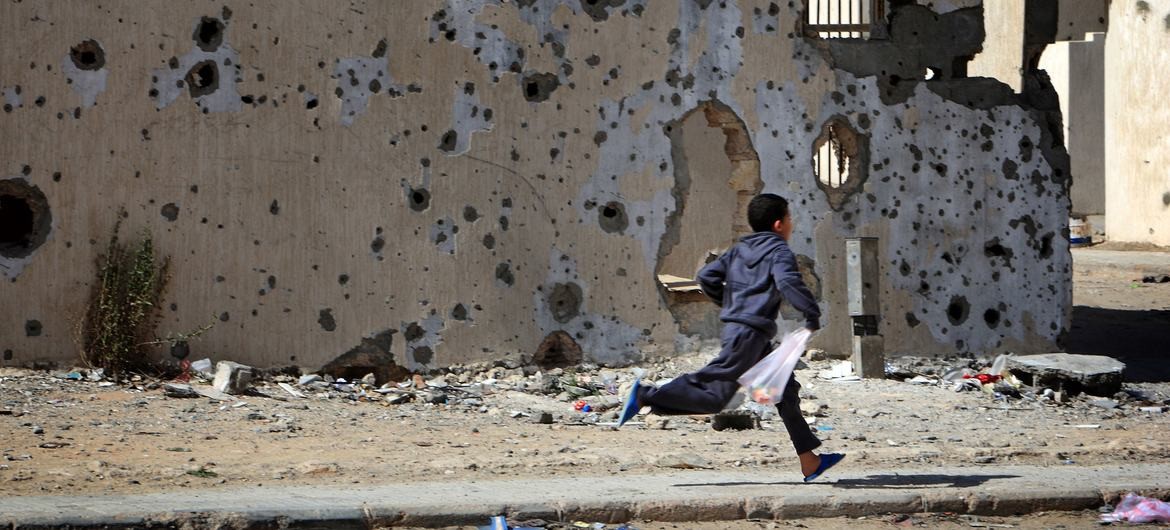 Một cậu bé chạy ngang qua những tòa nhà bị hư hại, bong tróc do đạn pháo trong cuộc xung đột, trên đường đi mua sắm về nhà ở thành phố Sirte.. (Nguồn: UNICEF)