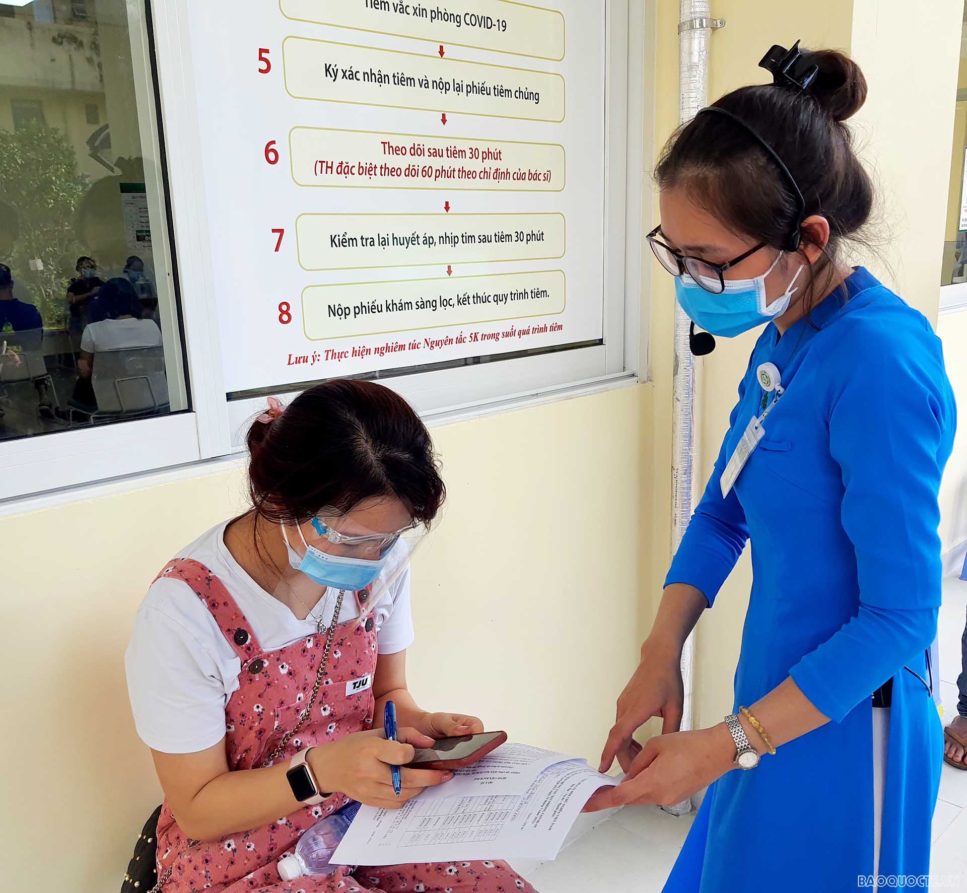 Nhân viên y tế trong bộ áo dài đang hướng dẫn người dân đăng ký tờ khai y tế trên mạng và sự nghiêm túc của người dân ngồi đợi đến lượt được tiêm vaccine. (Ảnh: Minh Hòa)